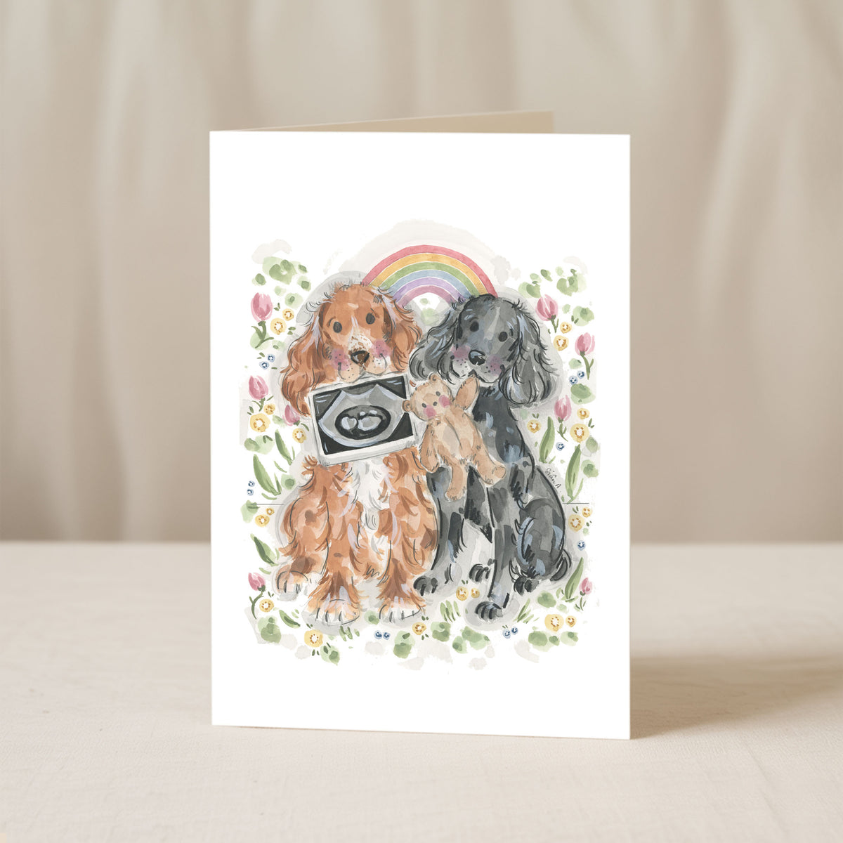 A white greeting card standing upright on a light beige surface, featuring a watercolour illustration of two dogs, a small baby doll, a rainbow, and flowers with a baby ultrasound scan held by the dog by sian alexia paine